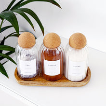 Load image into Gallery viewer, Three glass containers with cork lids on a wooden tray, with a plant in the background.