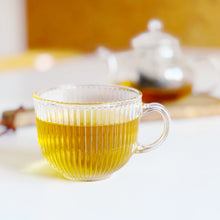 Load image into Gallery viewer, A clear ribbed glass teacup filled with golden yellow tea, set on a white surface with a softly blurred glass teapot and warm yellow background.