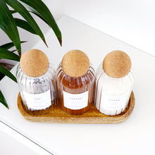 Load image into Gallery viewer, Three small glass bottles with cork stoppers on a wooden tray, with a plant in the background.
