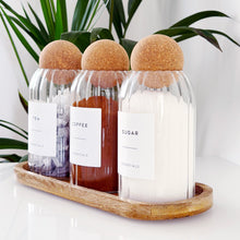 Load image into Gallery viewer, Three glass containers with cork lids on a wooden tray, with plants in the background.
